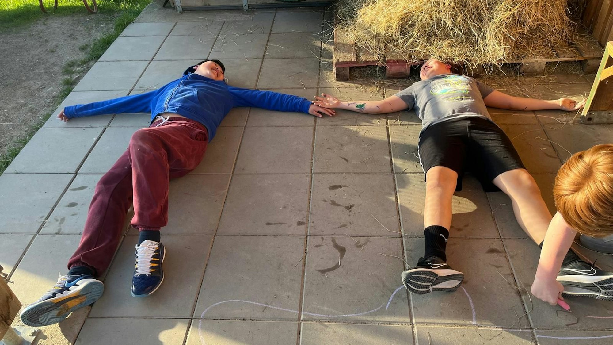 Zwei Kinder liegen auf dem Boden, sie lachen und halten sich an der Hand. Ein drittes malt mit Kreide einen Umriss ihrer Körper.