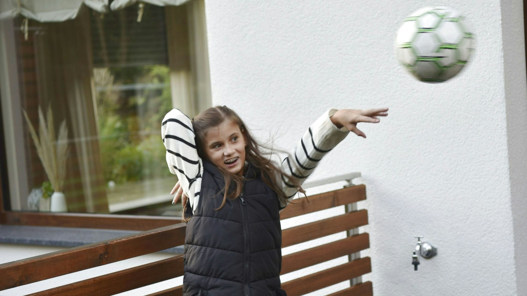 Josi Fischer steht auf der Terrasse ihres Elternhauses und wirft einen Ball. Sie trägt eine schwarze Weste, ein schwarz-weiß gestreiften Pulli und eine graue Jeans.