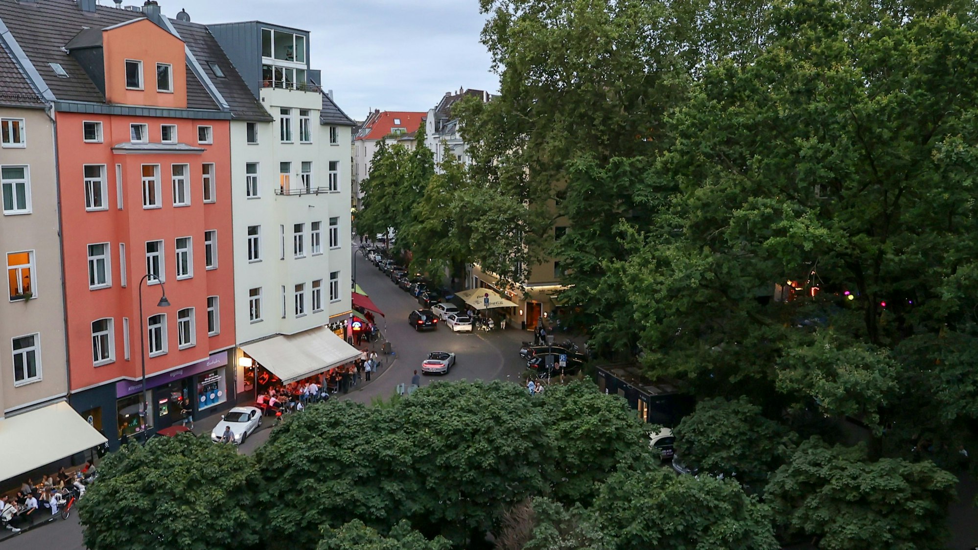 Zu sehen sind Häuser und Bäume am Brüsseler Platz in Köln und Menschen, die in Außenbereichen von Restaurants sitzen.