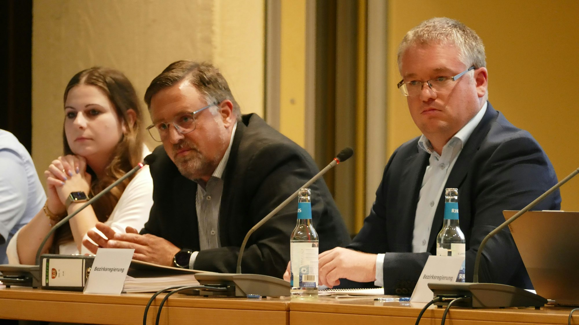 Eine Frau und zwei Männer sitzen an einem Tisch vor Mikrofonen.