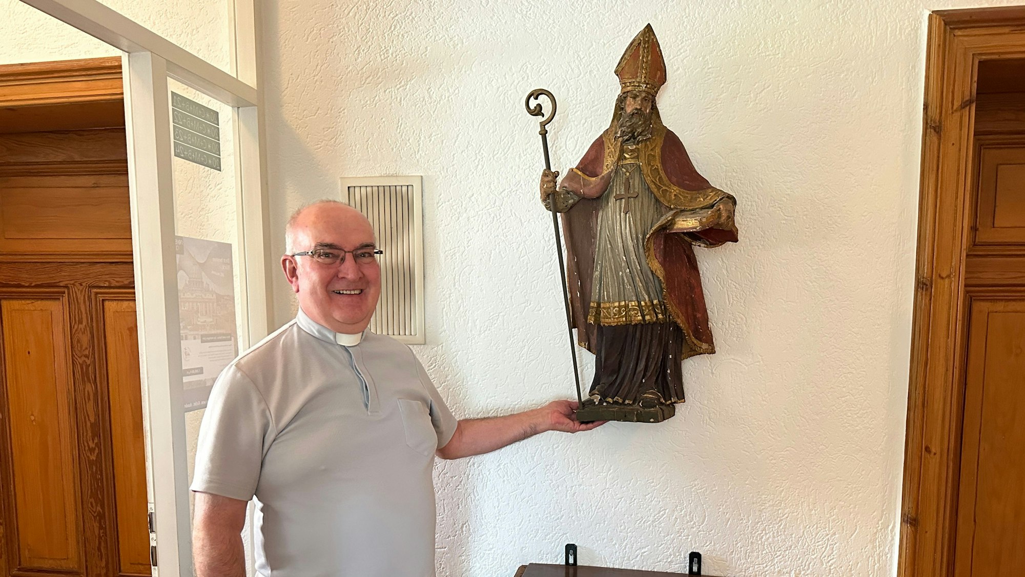Lambert Schäfer steht neben einer Heiligenfigur, er trägt ein hellgraues Hemd mit römischem Kragen.