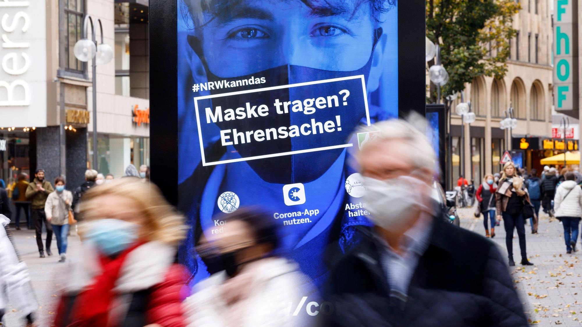 In Köln galt in der Fußgängerzone eine Maskenpflicht. Eine Straßenszene mit Passanten und Plakat mit dem Text "Maske tragen? Ehrensache" bilden das ab.