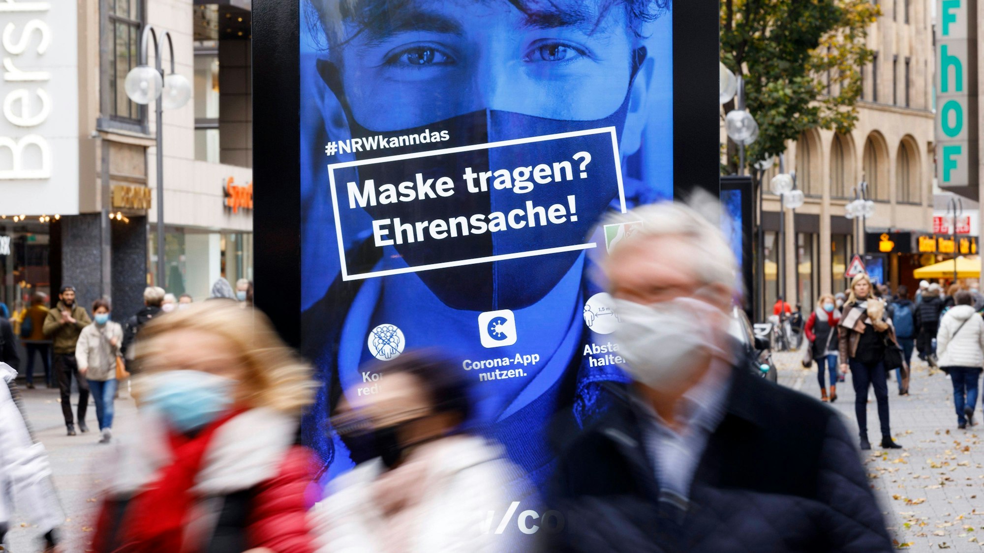 In Köln galt in der Fußgängerzone eine Maskenpflicht. Eine Straßenszene mit Passanten und Plakat mit dem Text "Maske tragen? Ehrensache" bilden das ab.