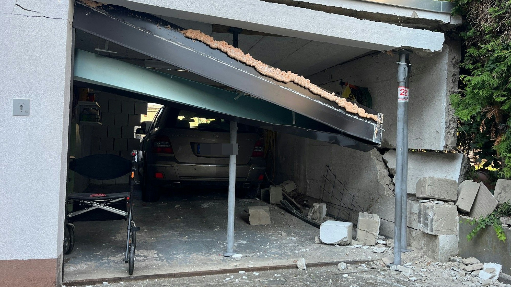 Eine Garage, deren Seitenwand eingebrochen ist. Mit Stützen wird die Decke gehalten. Ein Auto steht halb in der Garage, halb in der durchbrochenen Rückwand.