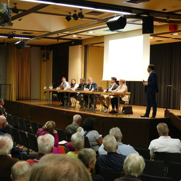 Besucher einer Informationsveranstaltung sitzen in einer Schulaula. Auf der Bühne sitzen Männer und Frauen an einem Podium.