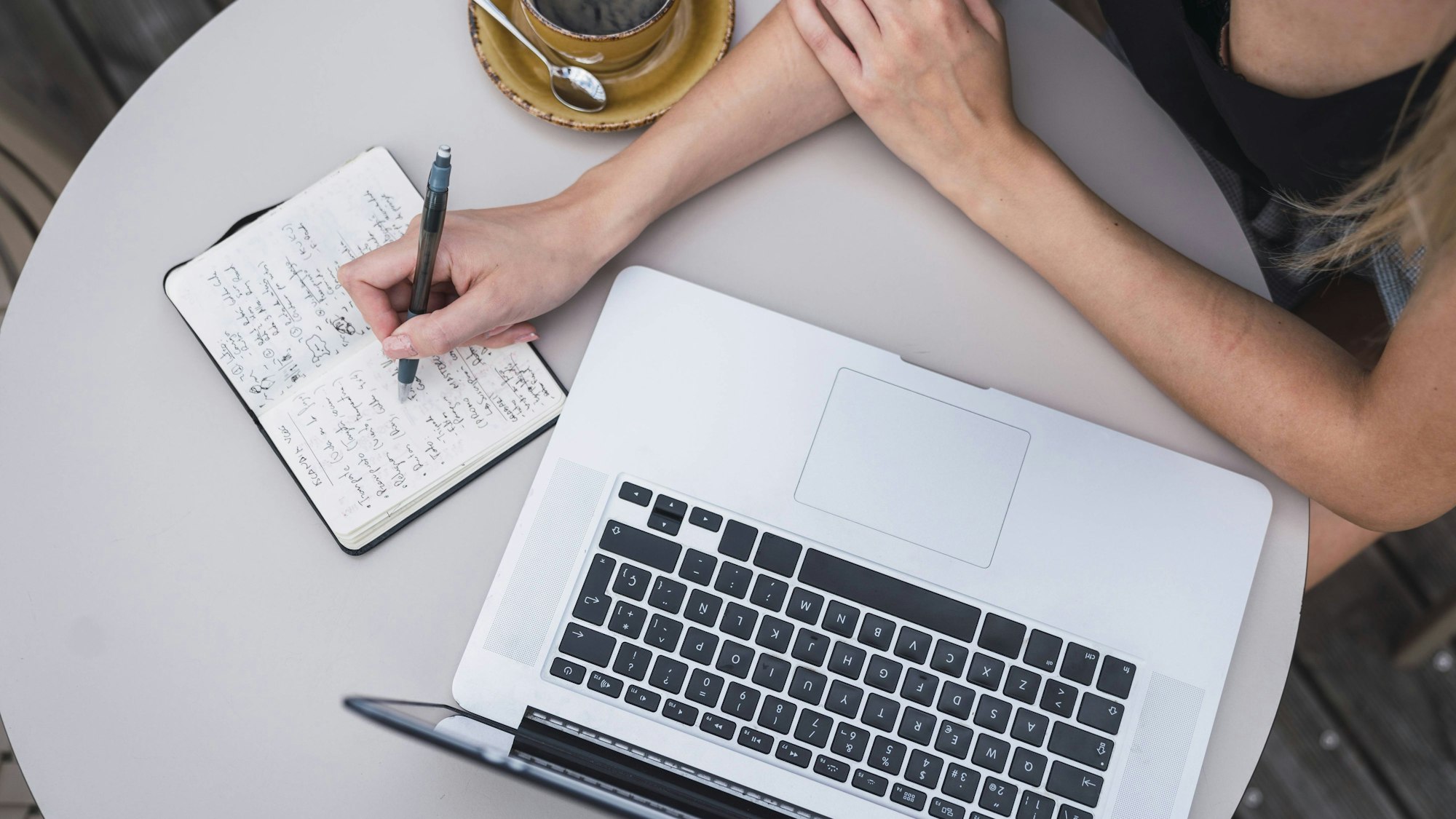 Eine Frau sitzt mit Kaffee an einem Tisch. Vor ihr steht ein Laptop, neben ihr liegt ein Notizbuch, in das sie mit einem Kugelschreiber schreibt.