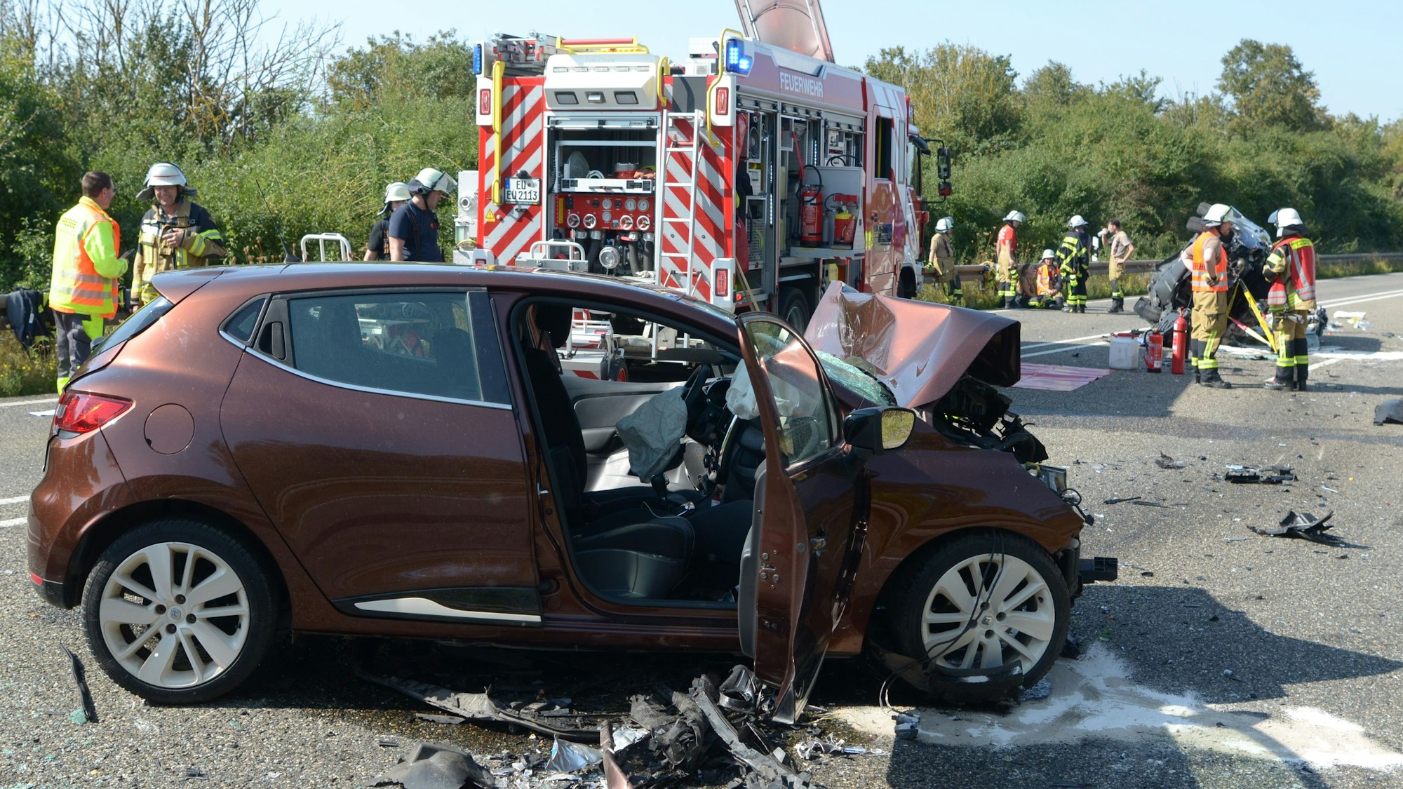 Das Auto steht quer zur Fahrtrichtung auf der Fahrbahn, im Hintergrund stehen ein Feuerwehrfahrzeug und Feuerwehrleute.