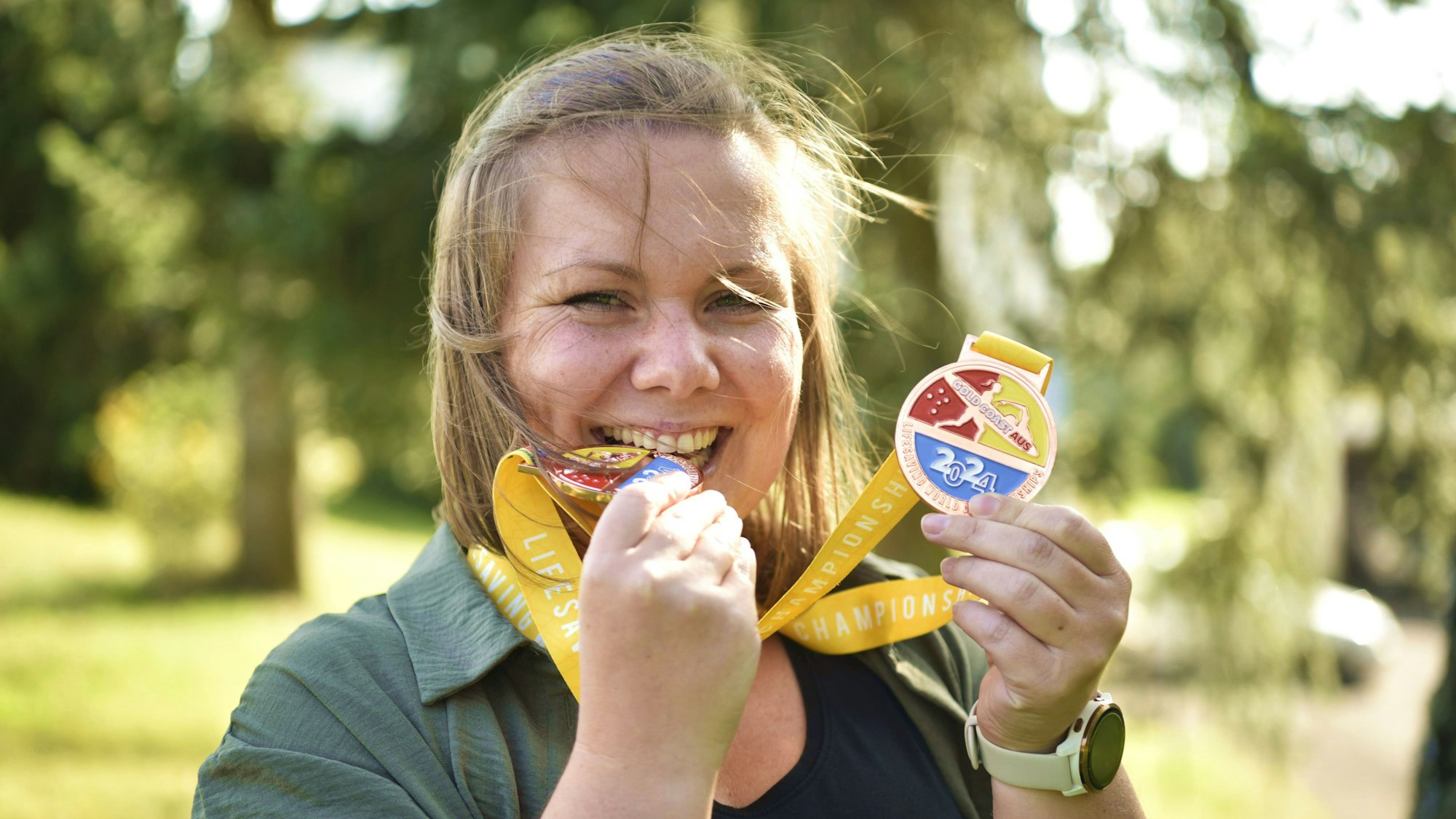 Eva Hoetgen beißt auf eine ihrer Bronze-Medaillen von den Weltmeisterschaften im Rettungsschwimmen 2024. Sie trägt schwarzes T-Shirt und grüne Bluse, im Hintergrund stehen Bäume.