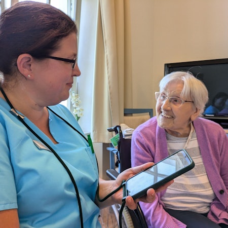 Jennifer Greuel sitzt bei einer Bewohnerin und hält das Handy vor sich, um die Daten und die Befindlichkeit der alten Dame zu dokumentieren.
