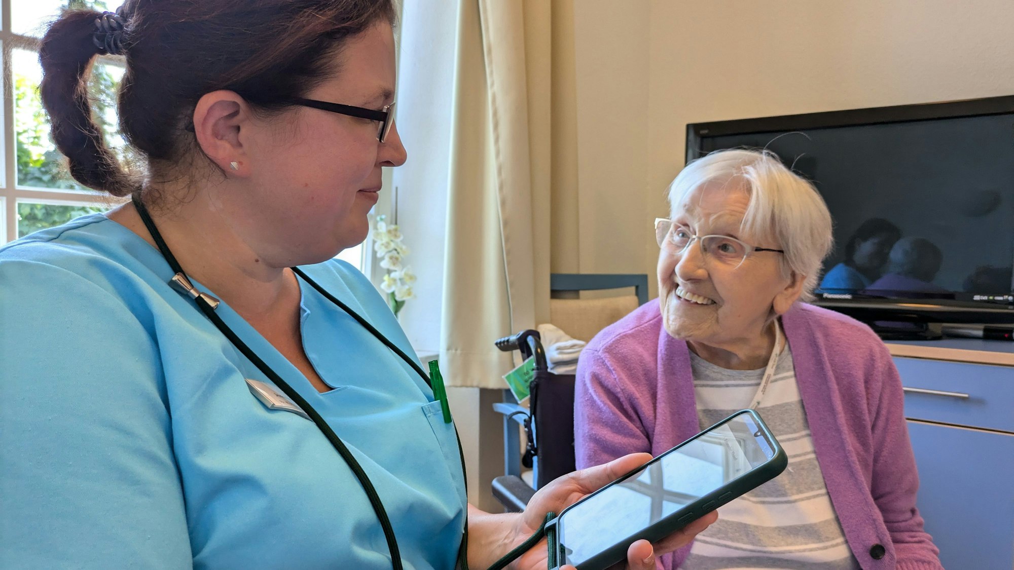 Jennifer Greuel sitzt bei einer Bewohnerin und hält das Handy vor sich, um die Daten und die Befindlichkeit der alten Dame zu dokumentieren.