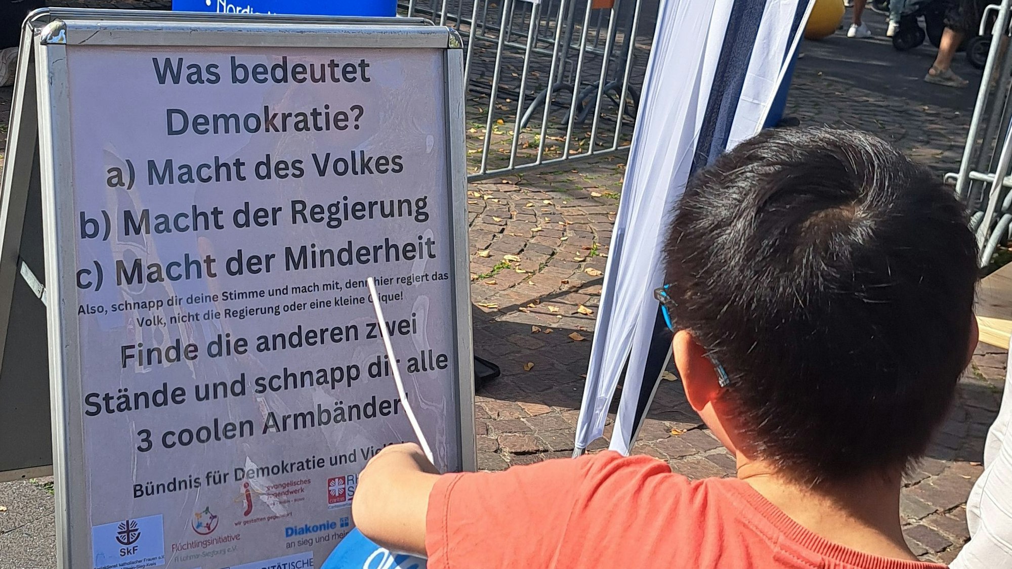 Ein Kind steht vor einem Plakat, auf dem nach der Bedeutung des Wortes Demokratie gefragt wird.