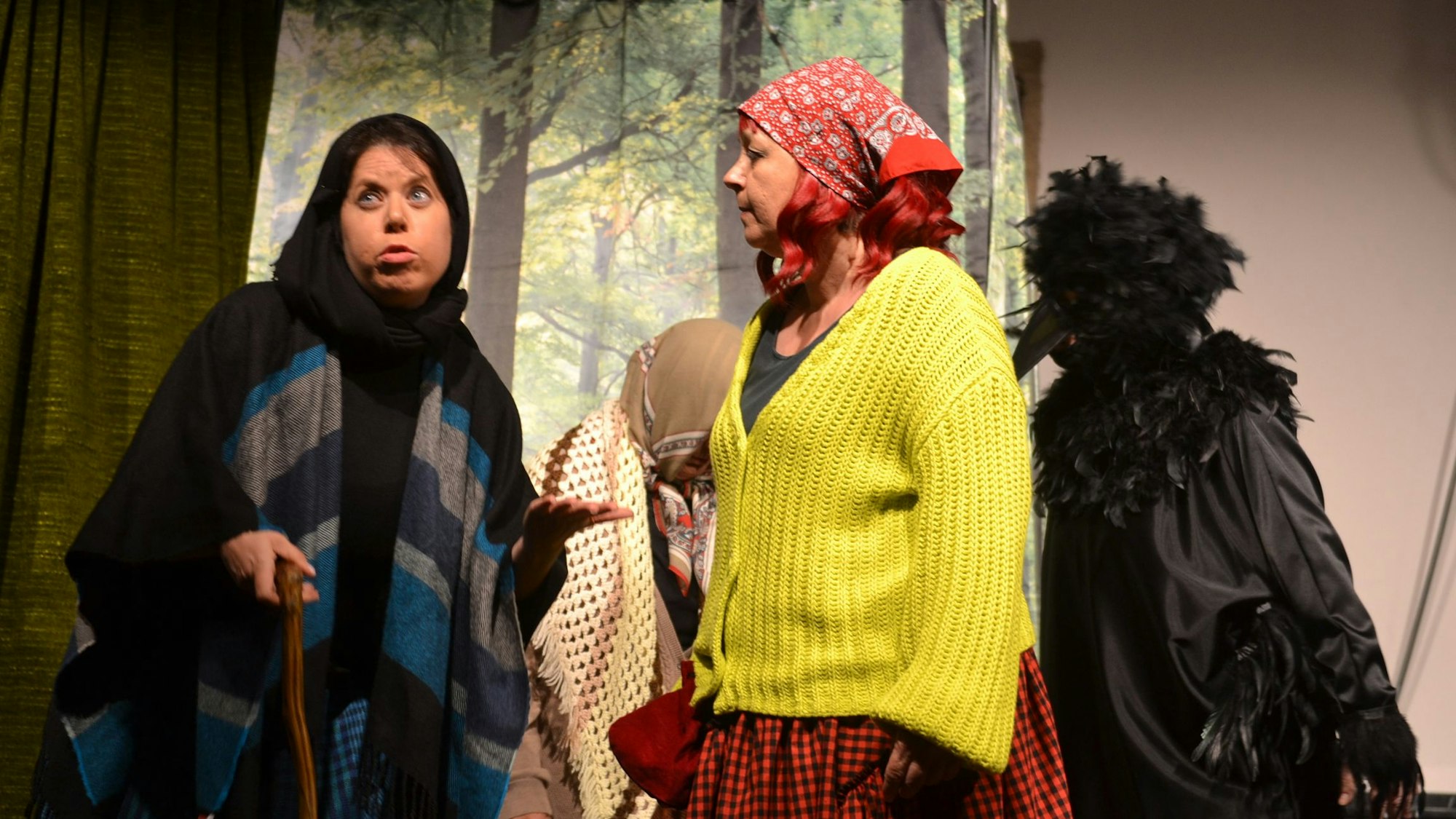 Zwei Schauspielerinnen mit Kopftüchern stehen auf der Bühne. Eine hält einen Stock in der Hand.