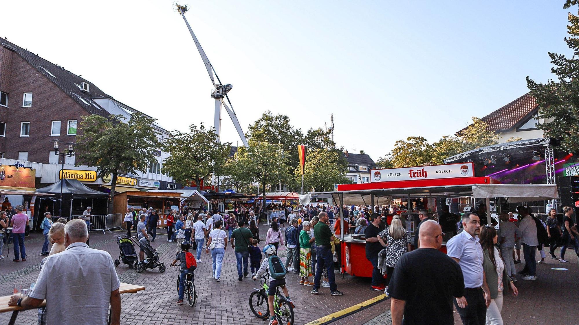 Menschen gehen über einen Platz mit Ständen, im Hintergrund ein Fahrgeschäft.