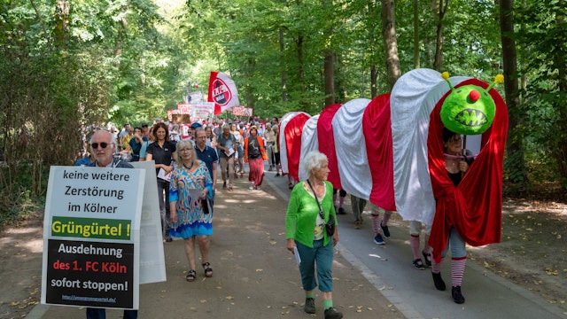 22.09.2024, Köln: Ultras des 1. FC Köln stören eine Demonstration der Bürgerinitivate „Grüngürtel für Alle“. Diese protestiert gegen den Bau des FC-Leistungszentrums im Grüngürtel. Eine rot-weiße Raupe Nimmersatt, symbolisch für das Anfressen des Grüngürtels, besucht die Orte, die der 1. FC Köln für seine Erweiterung betrachtet.