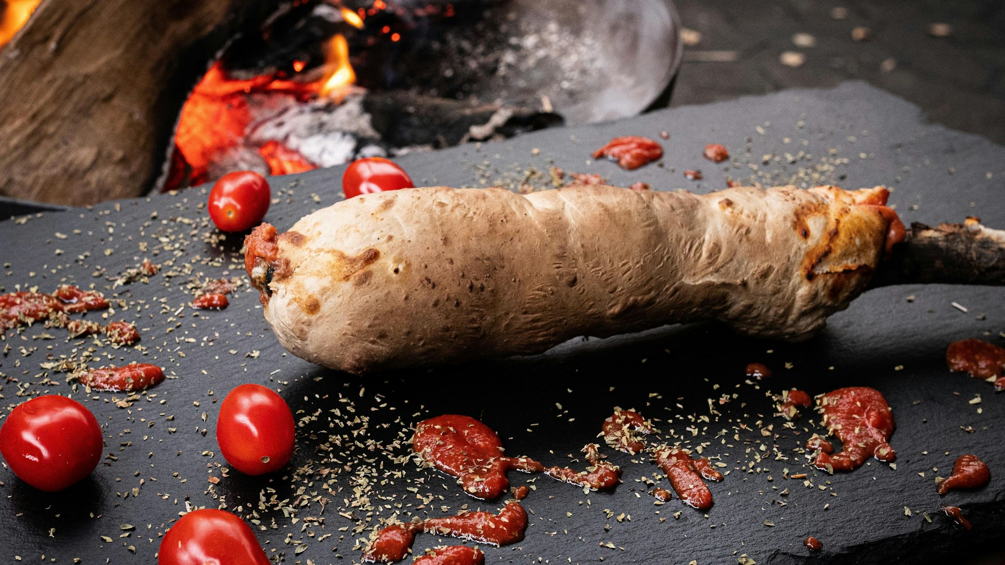 Mit Tomaten und Oregano vermischter Stockbrot-Teig backt am Feuer.