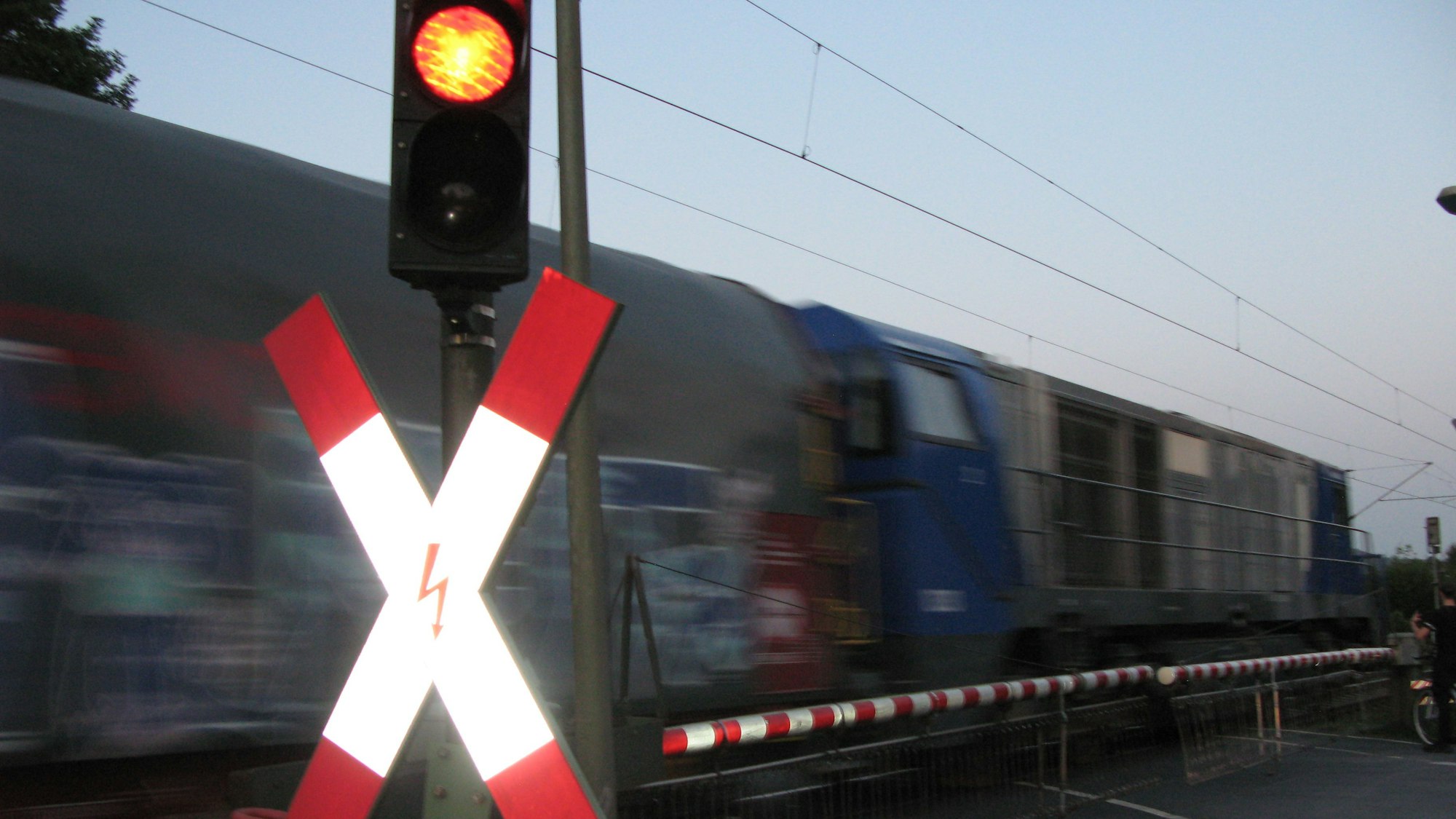 Ein Bahnübergang mit Andreaskreuz, Rotlicht und geschlossener Schranke, hinter der ein Güterzug fährt.