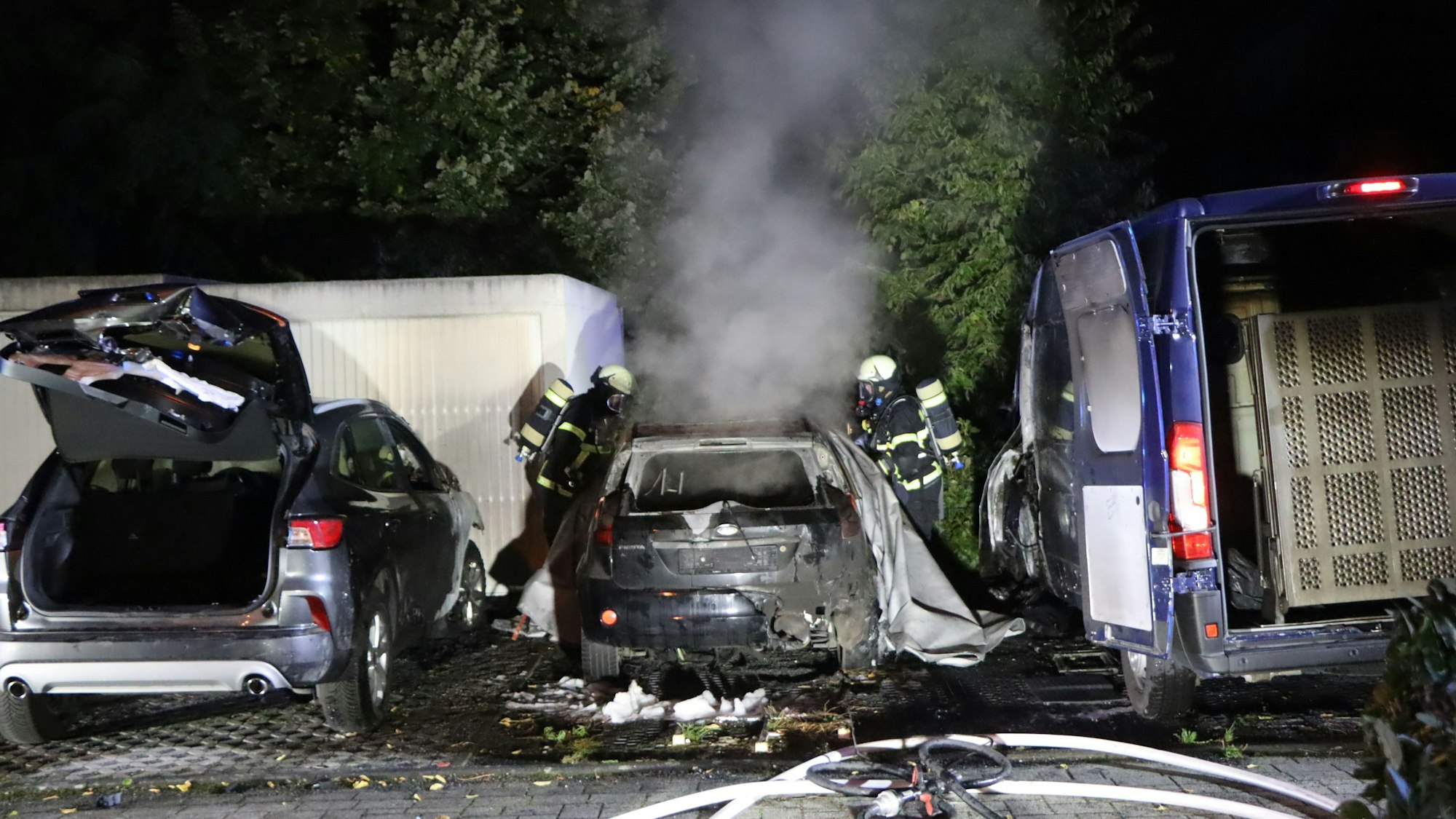 Drei Autos mit geöffnetem Kofferraum und Brandspuren. Das mittlere Fahrzeug raucht. Rechts und links stehen Feuerwehrleute.