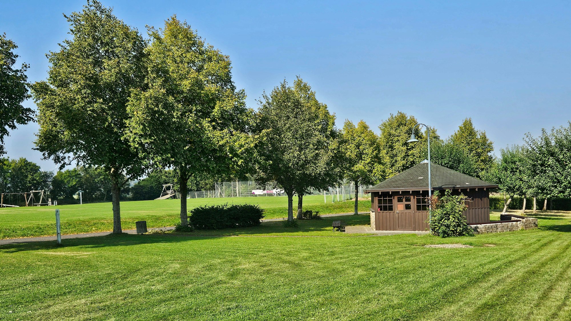 Blick auf die Wiese mit der Grillhütte und dem Sportplatz im Hintergrund.