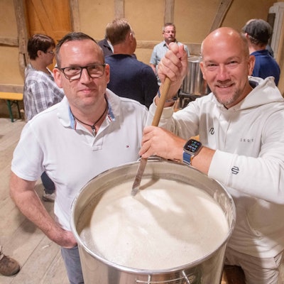 Das Foto zeigt zwei Männer, die in einem großen Topf mit Maische rühren.