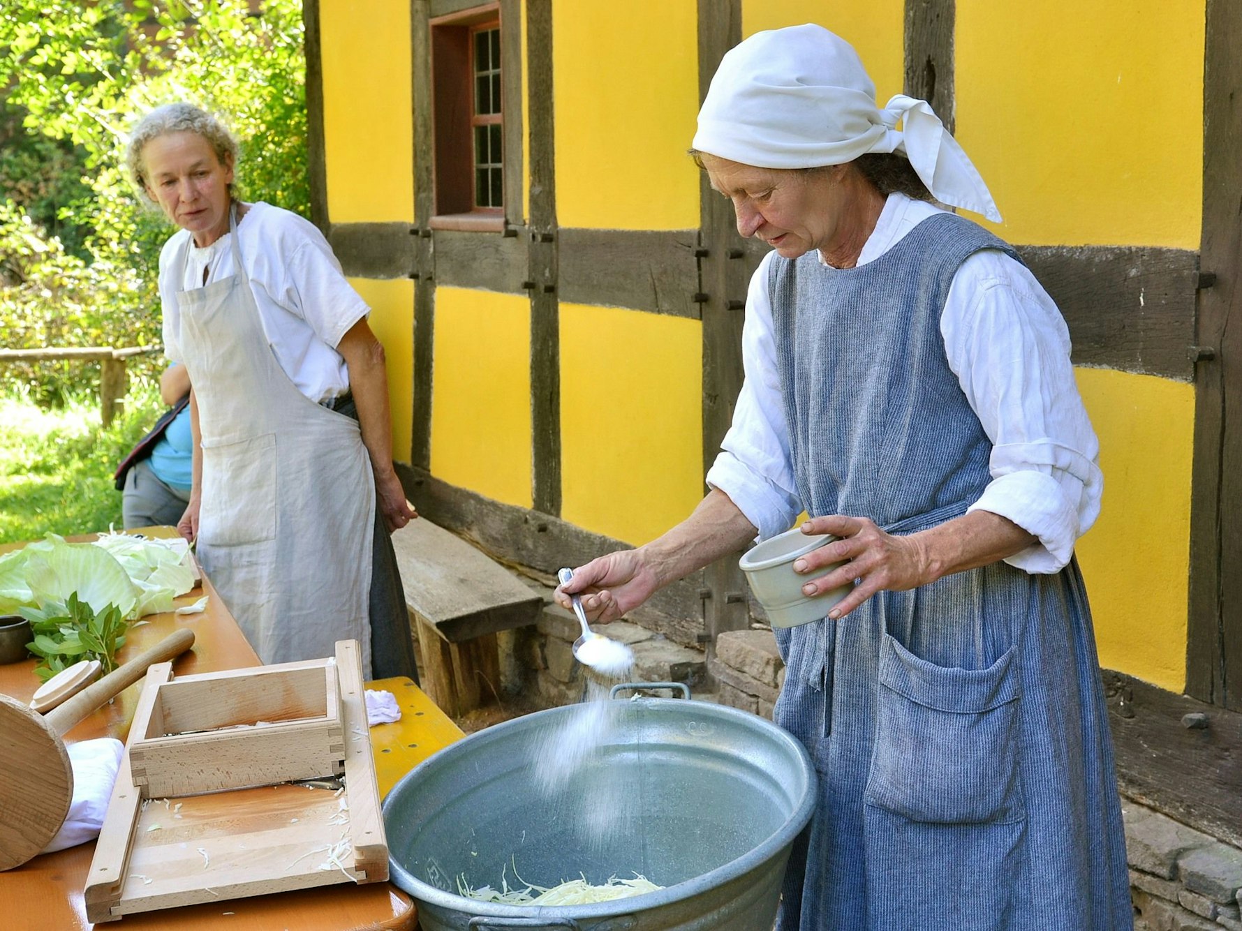 Von zwei Frauen mit Kochschürzen schüttet eine einen gehäuften Löffen Salz auf Sauerkraut in einem Topf.