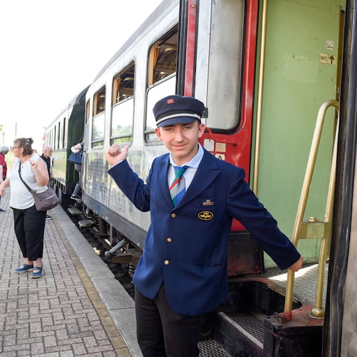 Ein Mann steht in der Originaluniform eines DB-Prüfers aus den 1960er Jahren vor einem Zug.