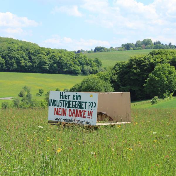 Eine Holztafel mit der Aufschrift Hier ein Industriegebiet? Nein danke! steht in einer hügeligen Wiesenlandschaft mit Bäumen im Hintergrund.