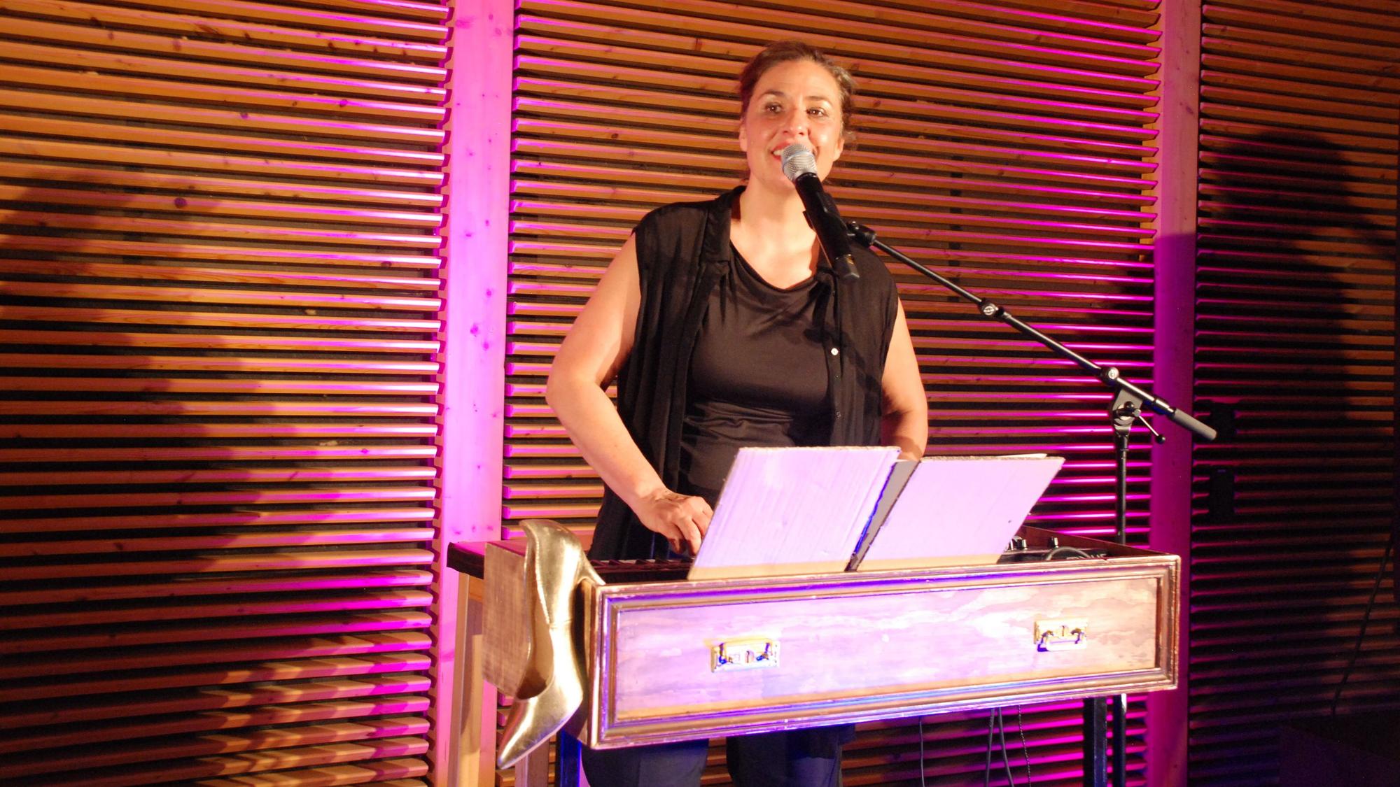 Kabarettistin Eva Eiselt steht auf einer Bühne vor einer Kommode, in die ein Bedienungsfeld eingebaut ist, und singt in ein Mikrofon.
