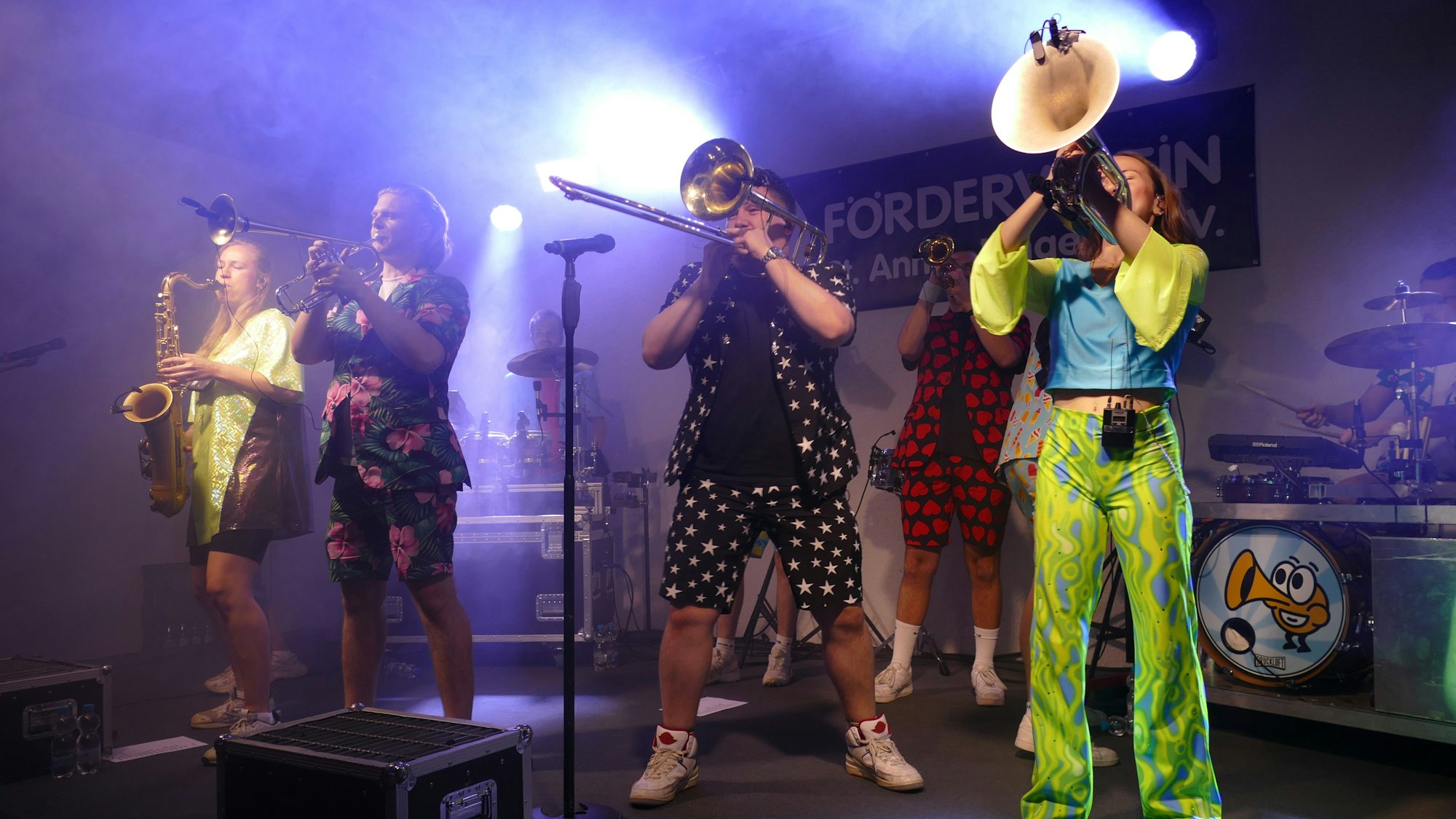 Männer und Frauen mit Blasinstrumenten und in bunter Kleidung stehen auf der Bühne.