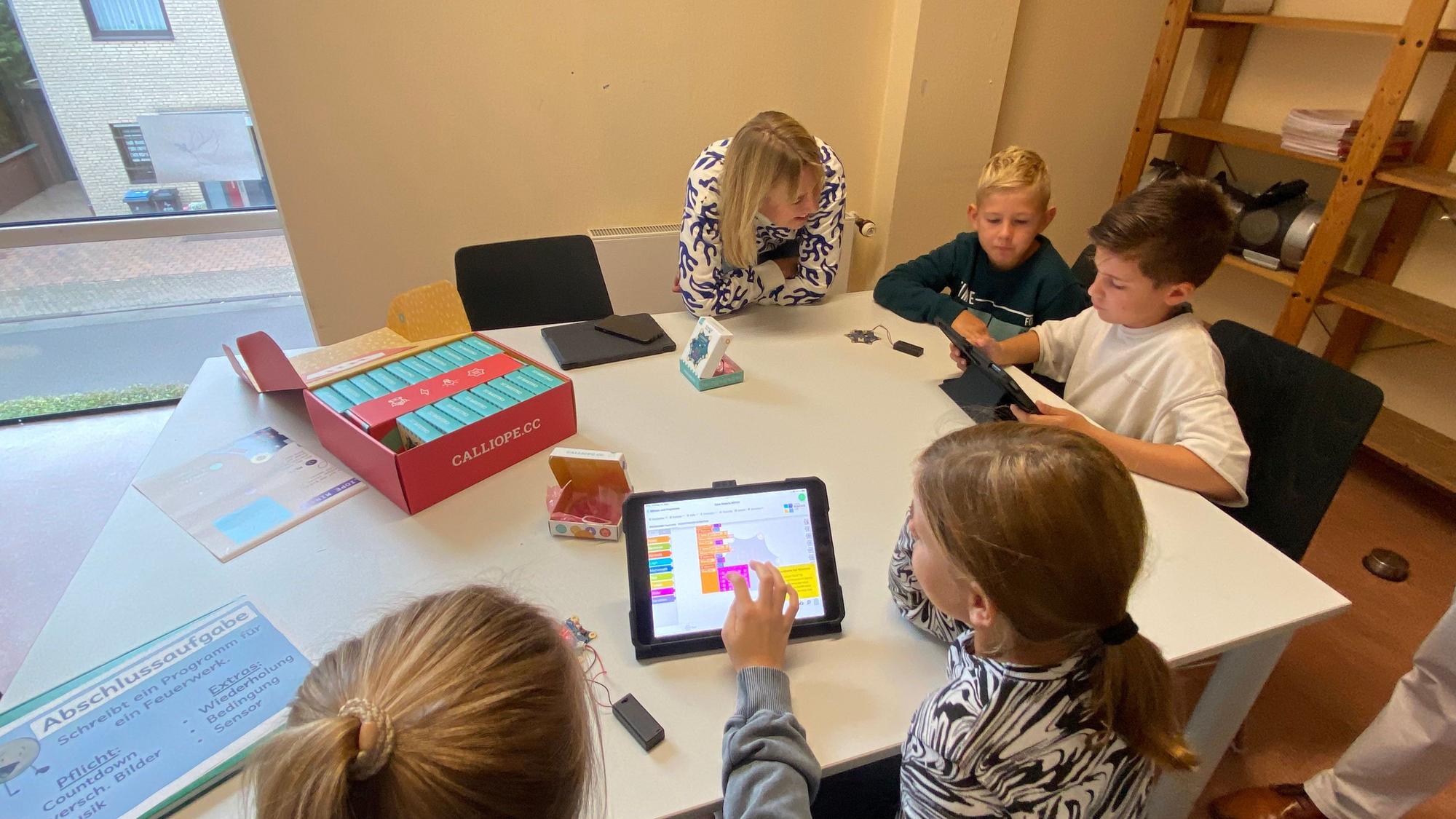 Kinder sitzen an einem Gruppentisch in der Schule. Eine Lehrerin beugt sich zu ihnen herunter.