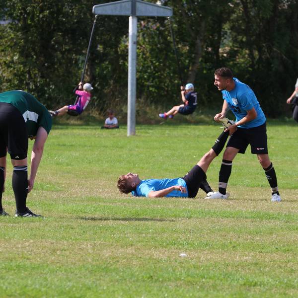Ein Fußballer liegt mit einem Krampf auf dem Sportplatz, ein Mitspieler hilft ihm. Ein Gegenspieler beugt sich nach vorne. Im Hintergrund gibt der Trainer das Zeichen zum Auswechseln.