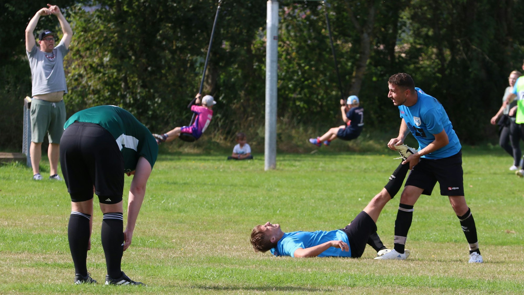 Ein Fußballer liegt mit einem Krampf auf dem Sportplatz, ein Mitspieler hilft ihm. Ein Gegenspieler beugt sich nach vorne. Im Hintergrund gibt der Trainer das Zeichen zum Auswechseln.