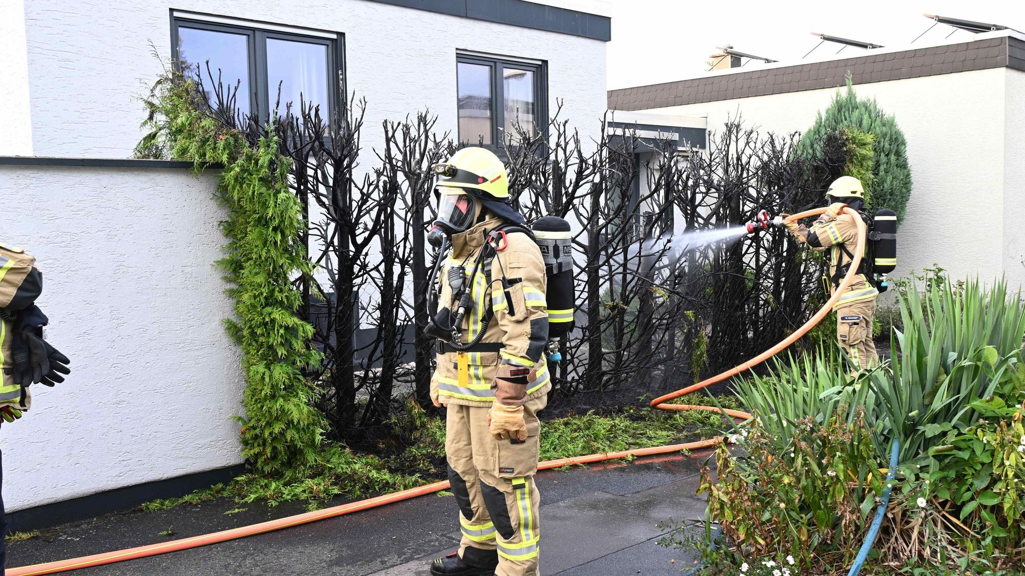 Feuerwehreinsatzkräfte vor einer verkohlten Hecke, im Hintergrund ein Wohnhaus.