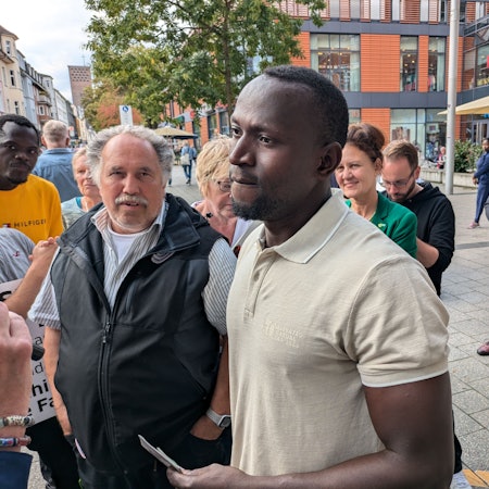 Ein junger und ein älterer Mann stehen mit anderen zusammen auf einem Platz.