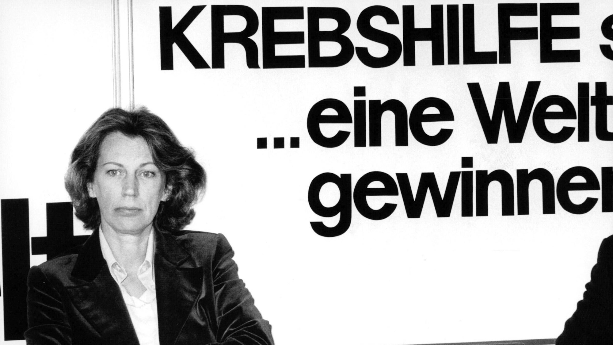 Mildred Scheel sitzt vor einem Plakat der Deutschen Krebshilfe