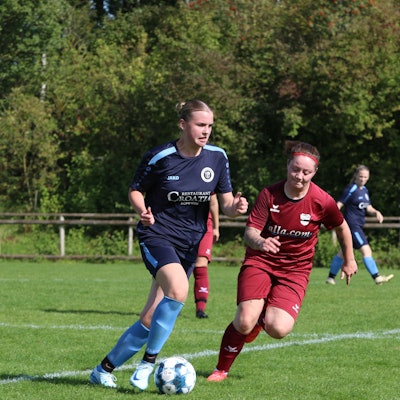 Eine Fußballerin im blauen Dress läuft mit einem Ball über den Platz. Ihr nähert sich eine Gegnerin im weinroten Trikot und mit Stirnband.