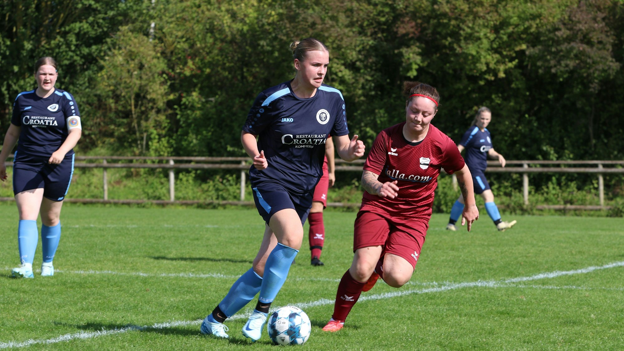 Eine Fußballerin im blauen Dress läuft mit einem Ball über den Platz. Ihr nähert sich eine Gegnerin im weinroten Trikot und mit Stirnband.