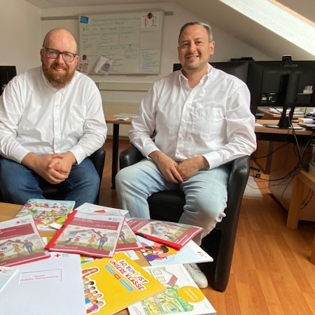 Zwei Männer sitzen vor einem Tisch, auf dem sämtliche mehrsprachige Broschüren der DRK-Integrationsagentur ausgelegt liegen.