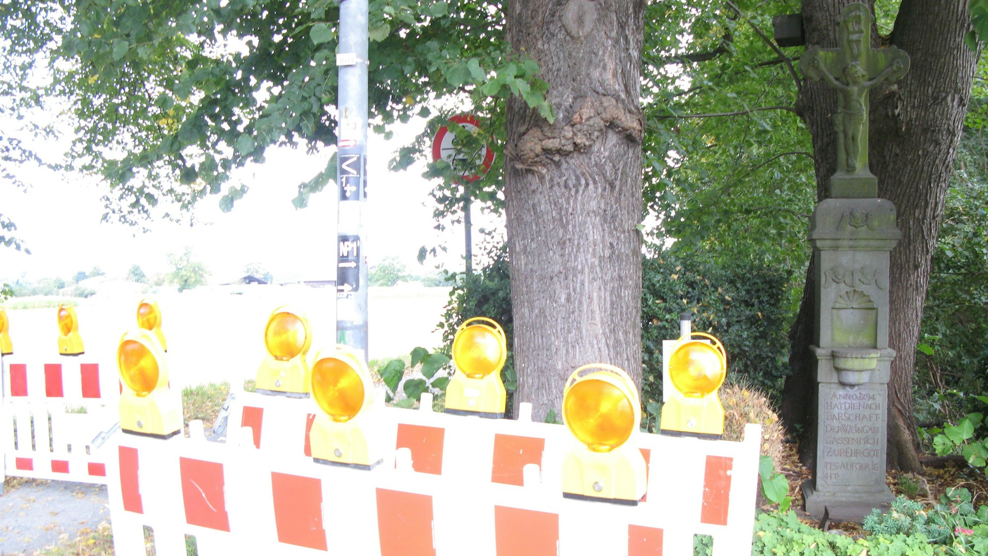 Rot-weiße Absperrelemente mit gelben Warnleuchten stehen auf dem Siegdamm an einer Baumgruippe mit steinernem Wegekreuz.