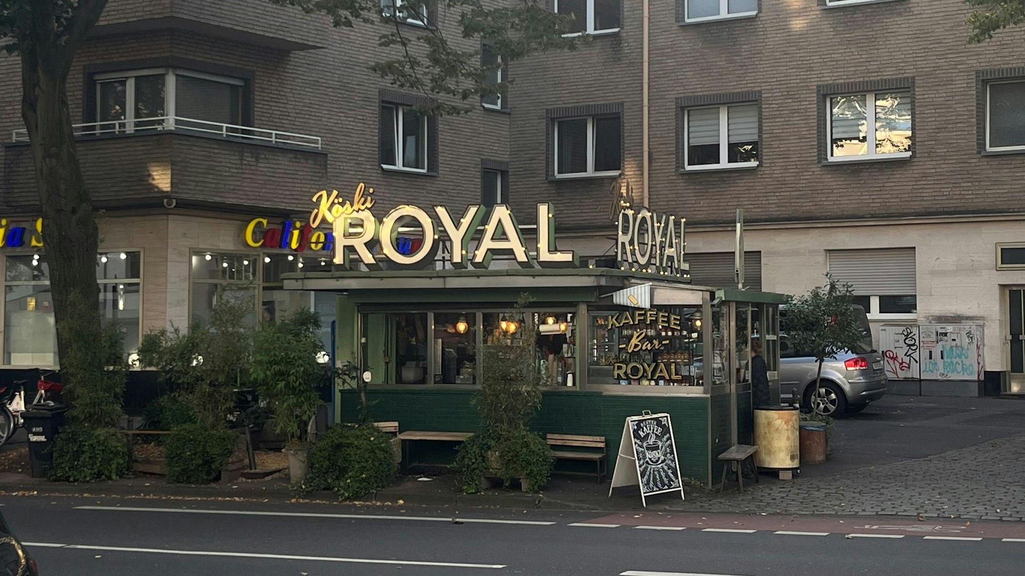 Das Köski Royal, ein Kaffeebüdchen in Köln-Braunsfeld, umgeben von Bäumen und einem Wohngebäude.