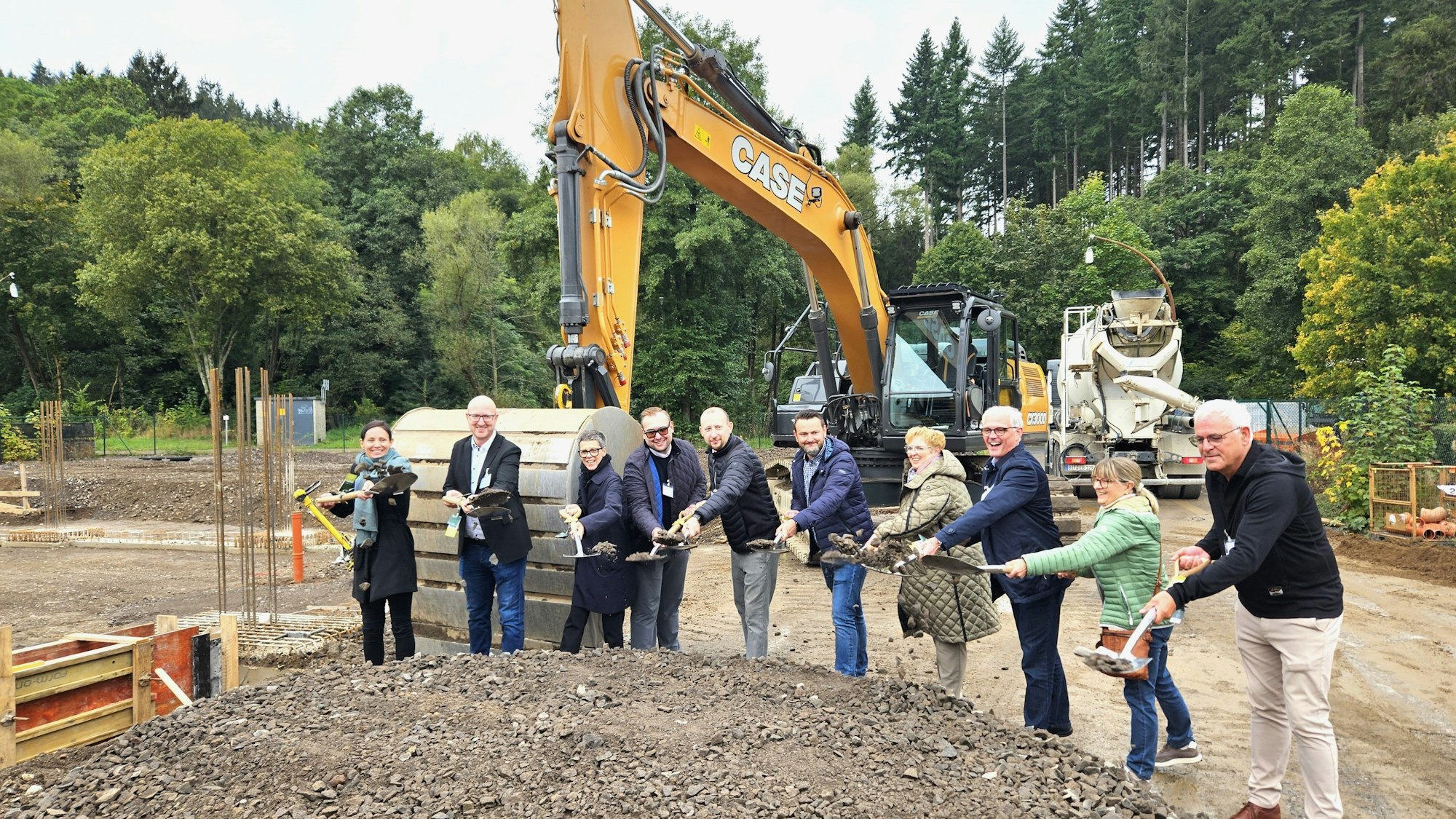 Vertreter der Unternehmen und aus Politik und Verwaltung mit Spaten in den Händen stehen vor einem Bagger.