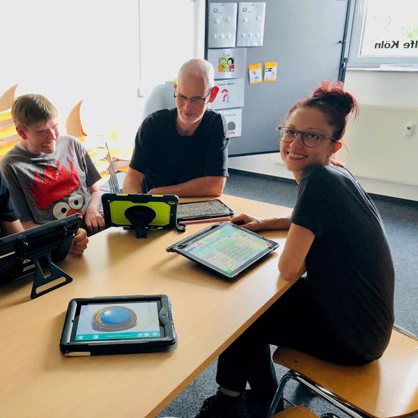 Zwei nichtsprechende junge Männer und zwei Mitarbeiter der Lebenshilfe Köln sitzen an einem Tisch vor ihnen liegen verschiedene Sprachcomputer, die auch Talker genannt werden.