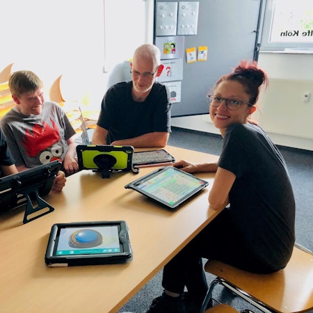 Zwei nichtsprechende junge Männer und zwei Mitarbeiter der Lebenshilfe Köln sitzen an einem Tisch vor ihnen liegen verschiedene Sprachcomputer, die auch Talker genannt werden.
