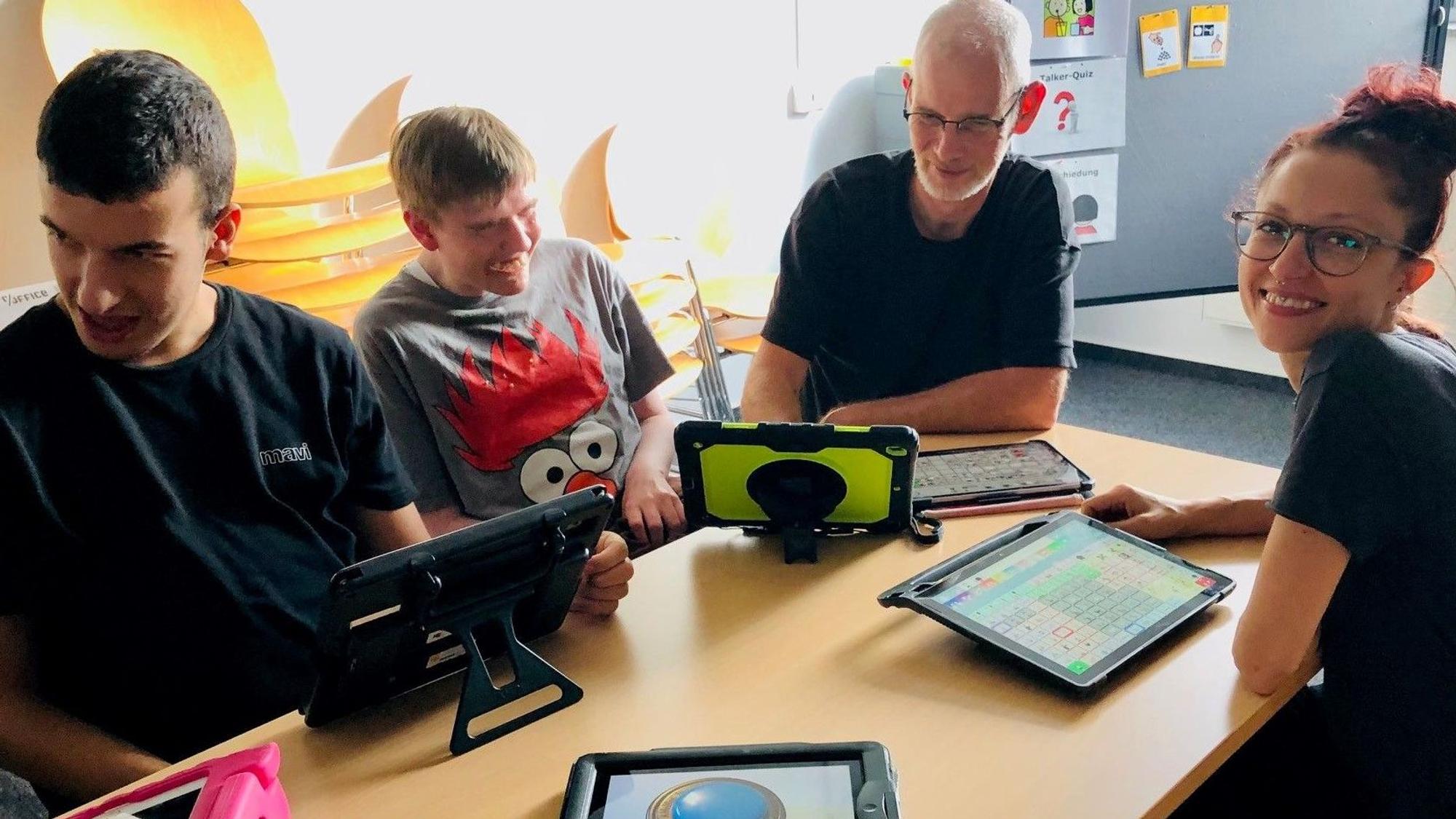 Zwei nichtsprechende junge Männer und zwei Mitarbeiter der Lebenshilfe Köln sitzen an einem Tisch vor ihnen liegen verschiedene Sprachcomputer, die auch Talker genannt werden.