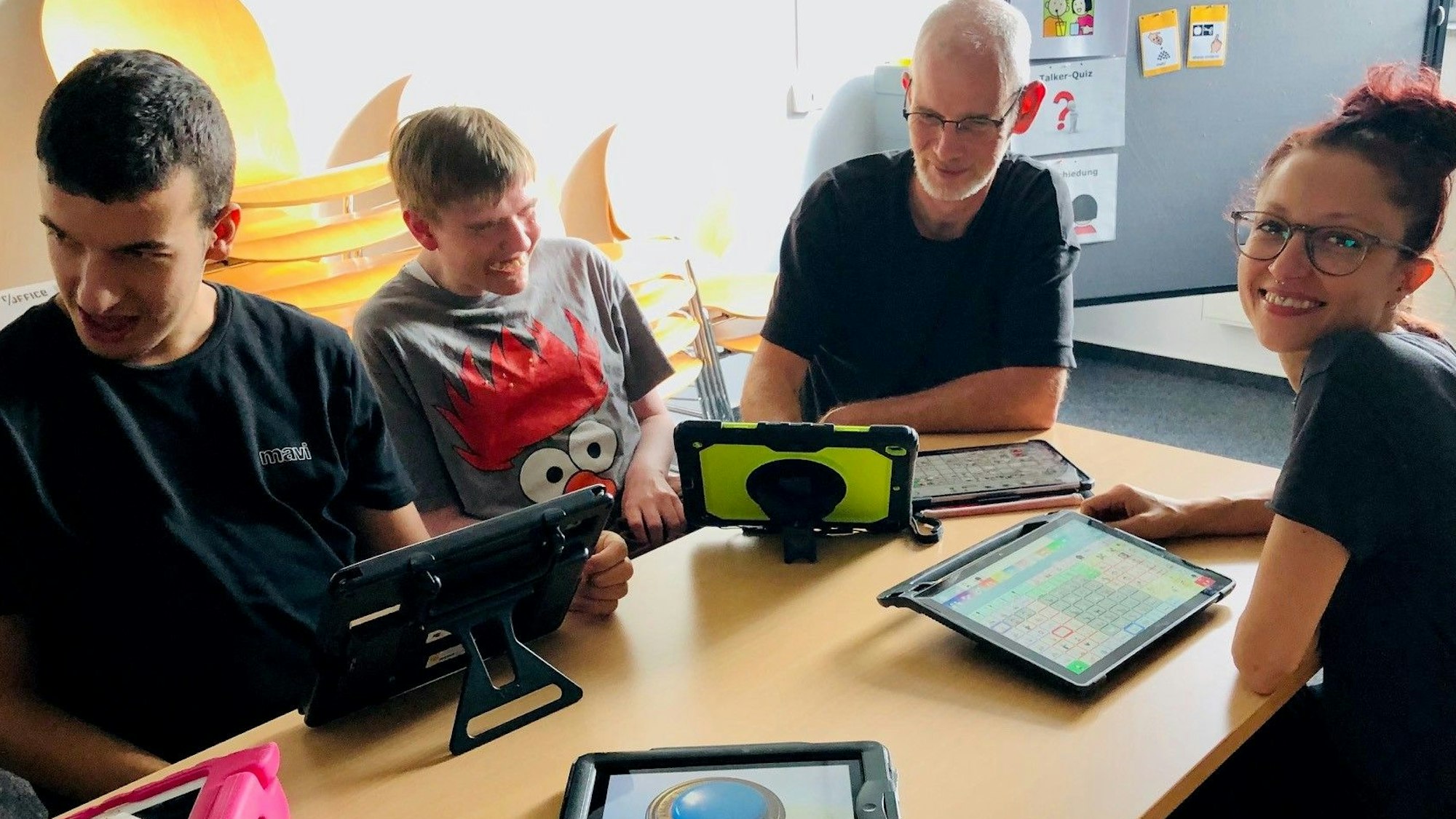 Zwei nichtsprechende junge Männer und zwei Mitarbeiter der Lebenshilfe Köln sitzen an einem Tisch vor ihnen liegen verschiedene Sprachcomputer, die auch Talker genannt werden.