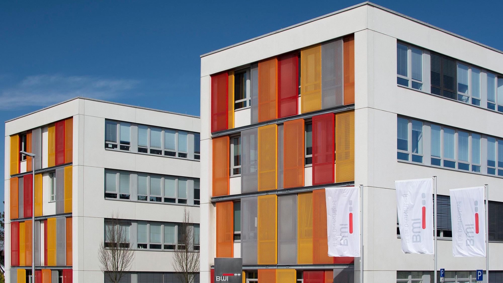 Ein Gebäude mit modernen Fensterläden in Orange, Grau und Rot. Davor wehen Fahnen mit dem BWI-Logo.