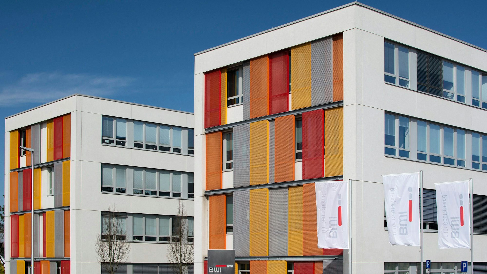 Ein Gebäude mit modernen Fensterläden in Orange, Grau und Rot. Davor wehen Fahnen mit dem BWI-Logo.