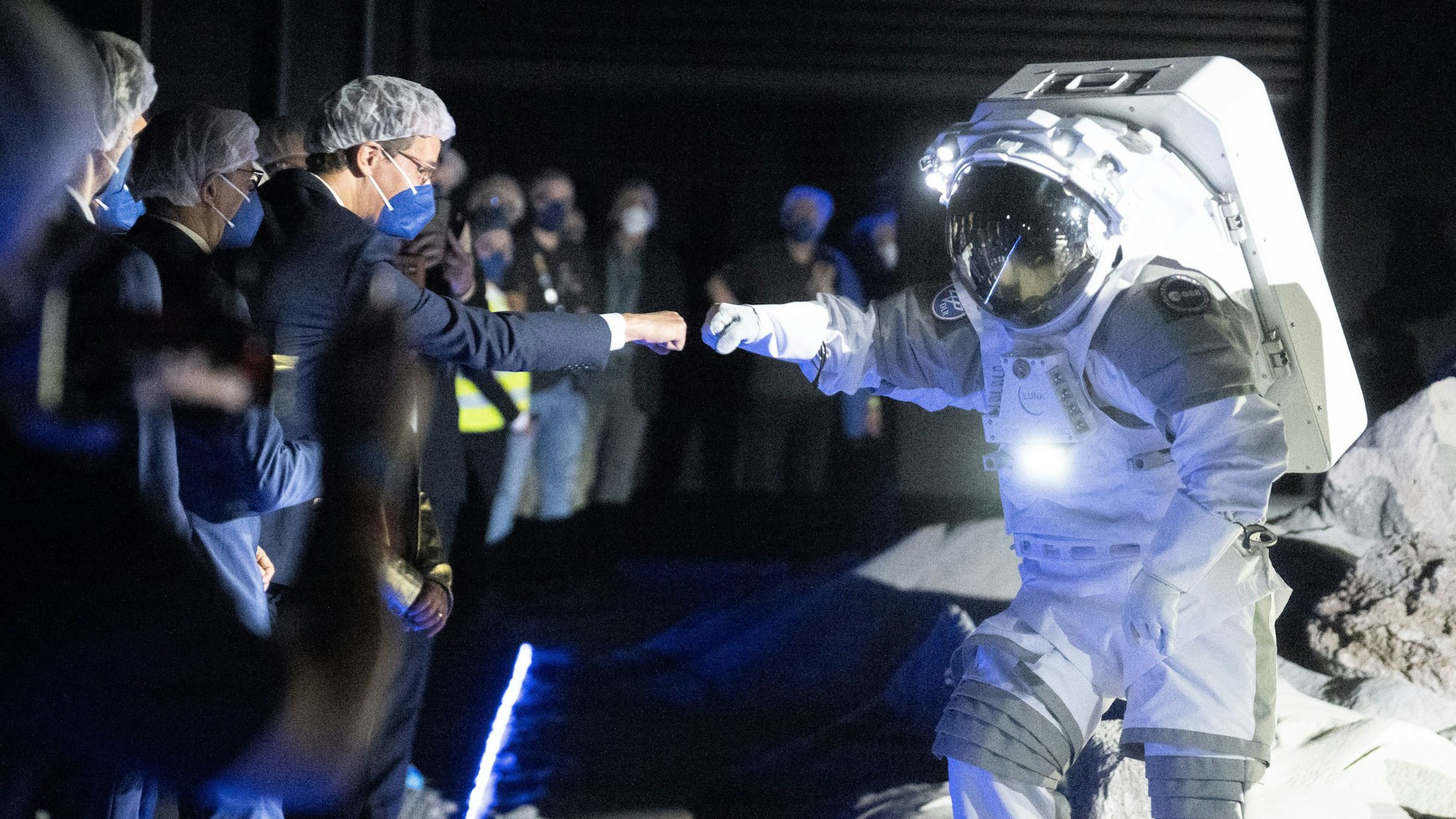 In einer Halle: Hendrik Wüst, der Ministerpräsident von Nordrhein-Westfalen, trägt eine OP-Haube und Maske und streckt einer Person im Raumfahrtanzug eine Faust zur Begrüßung entgegen.