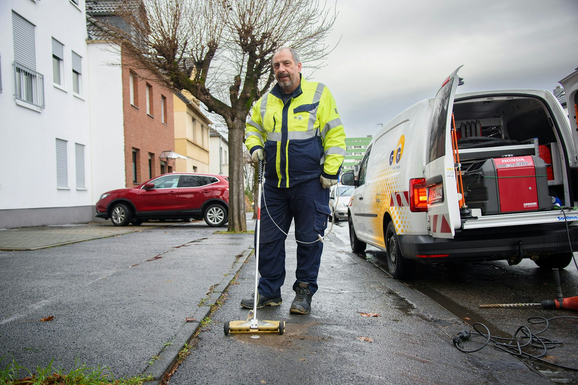 Ein Mann im Arbeitszug schiebt einen Detektor, der wie ein Staubsauger aussieht, vor sich her. Neben ihm ein Technikfahrzeug der GVG mit geöffneten Hecktüren.