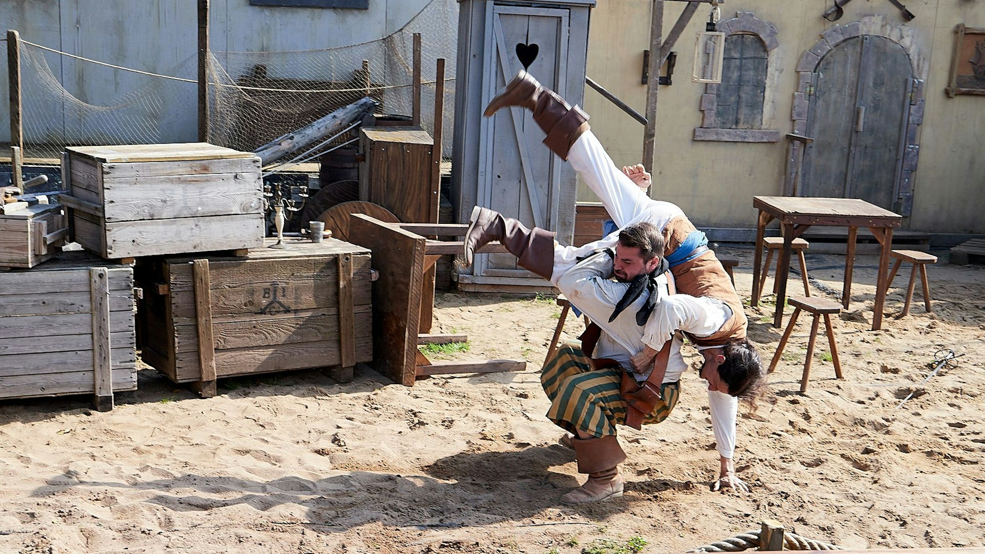 Ein als Pirat verkleideter Mann hockt auf sandigem Boden und wirft einen anderen als Pirat verkleideten Mann über seine Schulter. Im Hintergrund sind Holzkisten, ein Tisch, Stühle, ein Klohäuschen aus Holz und eine Straßenlaterne zu erkennen.