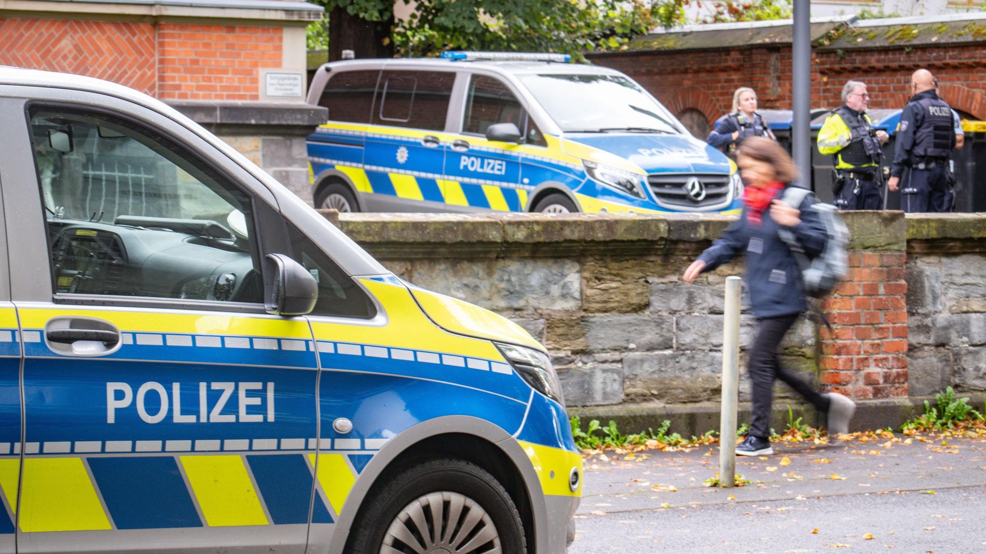 Polizisten stehen hinter einem Schild an einer Gesamtschule. Eine Person läuft vorbei.